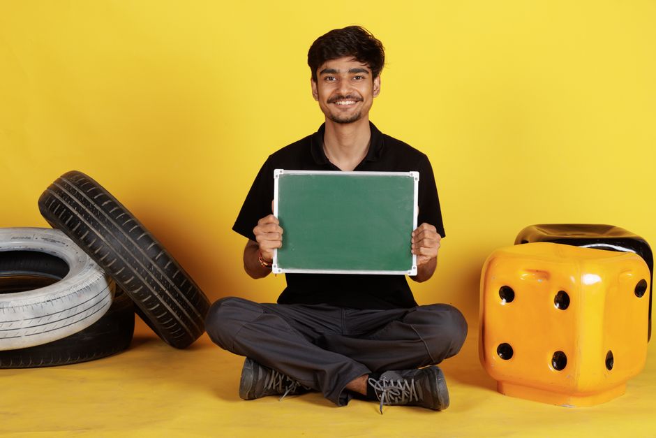 Smiling young man holding an empty green chalkboard amidst colorful props on a vibrant yellow backdrop.